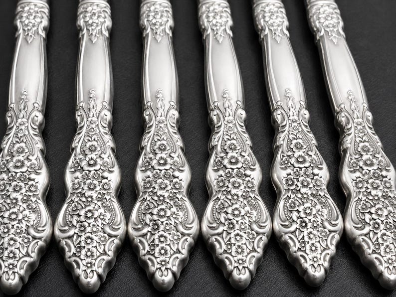 May include: A set of six silver-toned metal flatware handles arranged in a parallel row. Each handle features a detailed embossed floral pattern with small flowers and leaves extending from the base toward the center of the handle.