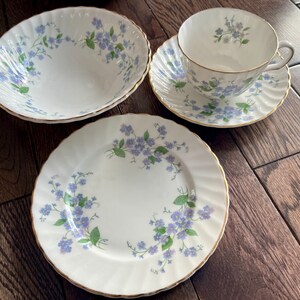 May include: A white porcelain teacup, saucer, bowl, and plate set. Each piece is decorated with a delicate blue floral pattern and gold trim. The set is arranged on a wooden surface, showcasing the elegant design.
