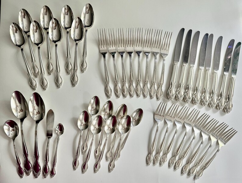 May include: A collection of vintage silverware, including spoons, forks, knives, and butter knives. The silverware is silver-toned with ornate floral detailing on the handles. The spoons have a rounded bowl shape. The forks have four tines.