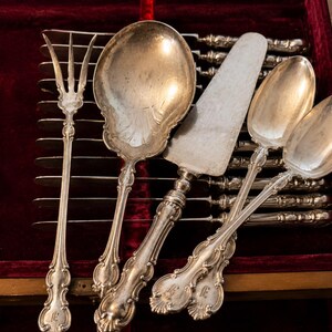 Puede incluir: Un surtido de utensilios de servicio de plata antiguos, que incluyen un tenedor, cucharas de servir y un servidor de pasteles, dispuestos en una caja de madera con un forro de terciopelo rojo. La cuberter&iacute;a presenta mangos ornamentados y un dise&ntilde;o cl&aacute;sico.