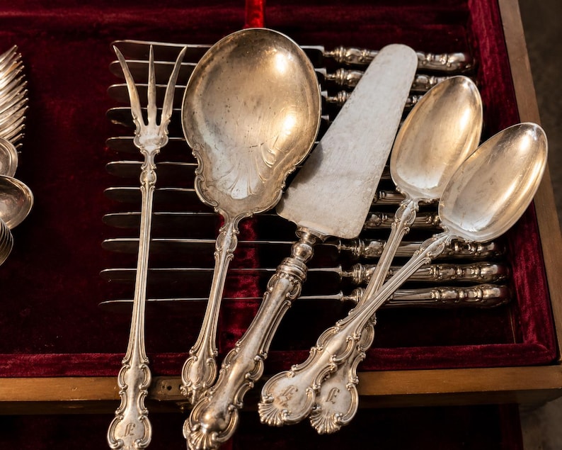 Puede incluir: Un surtido de utensilios de servicio de plata antiguos, que incluyen un tenedor, cucharas de servir y un servidor de pasteles, dispuestos en una caja de madera con un forro de terciopelo rojo. La cuberter&iacute;a presenta mangos ornamentados y un dise&ntilde;o cl&aacute;sico.