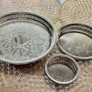 May include: Three silver-coloured serving trays of different sizes. The largest tray has an ornate, embossed floral design. The medium tray has a similar design, and the smallest is a simple, round tray. All have a decorative edge.