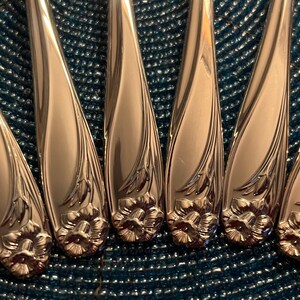 May include: Close-up of six polished silver spoons. Each spoon features a floral design at the base of the handle and a decorative pattern along the sides. The spoons are arranged on a textured blue surface, reflecting light.