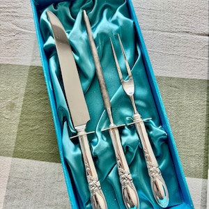 Op de afbeelding: Een zilveren vleeswarenset, inclusief mes, vork en aanzetstaal, gepresenteerd in een blauwe doos met turquoise satijnen voering. De handvatten hebben een bloemenmotief. Het mes is ongeveer 30 cm lang.