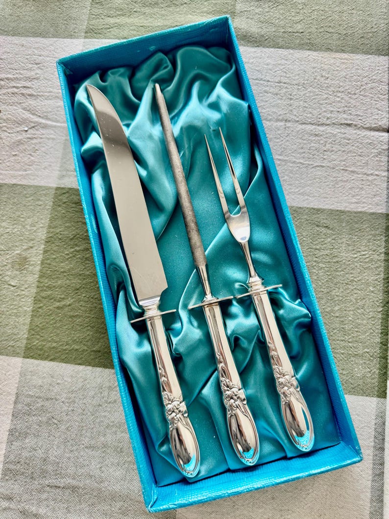 Op de afbeelding: Een zilveren vleeswarenset, inclusief mes, vork en aanzetstaal, gepresenteerd in een blauwe doos met turquoise satijnen voering. De handvatten hebben een bloemenmotief. Het mes is ongeveer 30 cm lang.