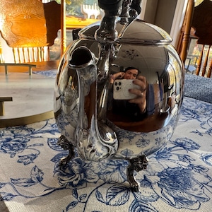 May include: A polished silver-tone metal teapot with a rounded body, a curved spout, and a lid. The teapot features ornate, detailed feet and a reflective surface that shows a distorted view of the surroundings.