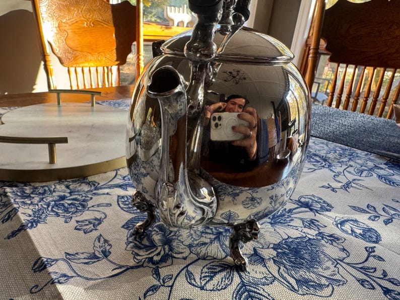 May include: A polished silver-tone metal teapot with a rounded body, a curved spout, and a lid. The teapot features ornate, detailed feet and a reflective surface that shows a distorted view of the surroundings.