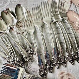 May include: A collection of ornate silver-colored flatware, including spoons and forks. The silverware features detailed floral and scrollwork designs on the handles. The spoons have a rounded bowl shape, and the forks have multiple tines. The arrangement suggests a formal dining setting.