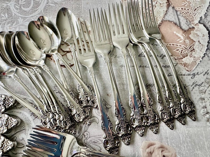May include: A collection of ornate silver-colored flatware, including spoons and forks. The silverware features detailed floral and scrollwork designs on the handles. The spoons have a rounded bowl shape, and the forks have multiple tines. The arrangement suggests a formal dining setting.