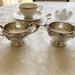 Puede incluir: Un juego de azucarero y lechera de plata con detalles florales en relieve en los laterales y las asas. El conjunto se acompa&ntilde;a de una taza de t&eacute; de porcelana blanca con borde dorado sobre un plato, una cuchara peque&ntilde;a y un cuenco de cristal.