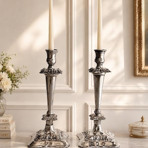 May include: A pair of tall, polished silver-toned metal candlesticks with ornate, embossed floral detailing on the square bases and central stems. Each candlestick holds a single, slender, off-white taper candle in the top socket.