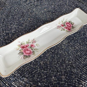 May include: A white ceramic serving dish with a gold trim and a floral design featuring pink roses. The dish is shaped like a long rectangle with rounded edges.