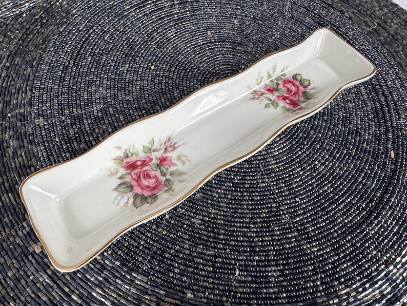 May include: A white ceramic serving dish with a gold trim and a floral design featuring pink roses. The dish is shaped like a long rectangle with rounded edges.