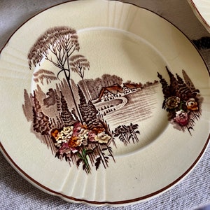 May include: A cream-coloured ceramic plate with a brown and white landscape design. The plate features a cottage, trees, and flowers.