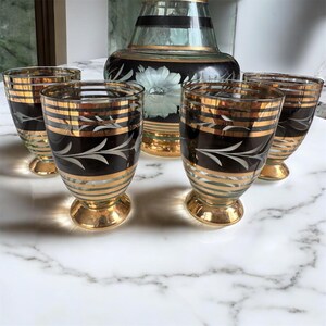 May include: Set of four clear glass shot glasses with gold and black accents. The glasses have a decorative etched design and are set on a white marble surface.