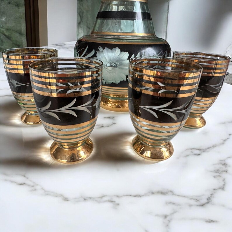 May include: Set of four clear glass shot glasses with gold and black accents. The glasses have a decorative etched design and are set on a white marble surface.