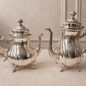 May include: Two polished silver teapots with ornate, swirling embossed patterns, curved handles, and decorative spouts. Each teapot features a matching lid with a small finial and stands on three detailed, curved feet on a light surface.