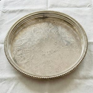 May include: A round silver-toned serving tray featuring an intricate embossed floral pattern across the center surface, a decorative pierced lattice border, and a twisted rope-style edge detail along the outer rim of the circular platter.
