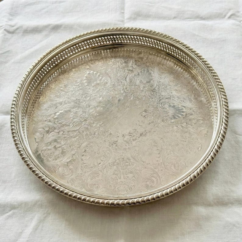 May include: A round silver-toned serving tray featuring an intricate embossed floral pattern across the center surface, a decorative pierced lattice border, and a twisted rope-style edge detail along the outer rim of the circular platter.