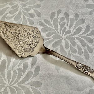 K&ouml;nnte beinhalten: Ein silberfarbener Tortenheber aus Metall mit einer verzierten dreieckigen Klinge, die ein eingraviertes Blumenmuster und ein zentrales Korbmotiv aufweist. Der Griff ist schlank mit einem Muscheldesign am Hals und einem abgerundeten Ende.