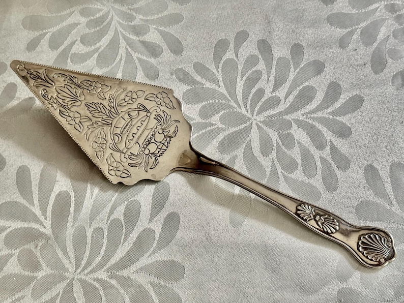 K&ouml;nnte beinhalten: Ein silberfarbener Tortenheber aus Metall mit einer verzierten dreieckigen Klinge, die ein eingraviertes Blumenmuster und ein zentrales Korbmotiv aufweist. Der Griff ist schlank mit einem Muscheldesign am Hals und einem abgerundeten Ende.