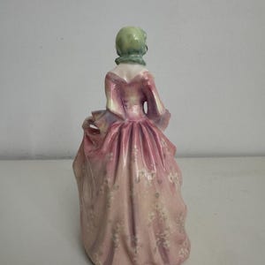 以下が含まれることがあります： 淡いピンク色のロングドレスを纏った人物の磁器製フィギュア。ドレスには繊細な白い花柄が施されている。人物は淡い緑色のヘッドカバーを着用しており、背後から見た姿で、ドレスのひだやドレープのディテールが表現されている。