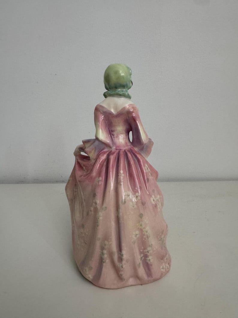以下が含まれることがあります： 淡いピンク色のロングドレスを纏った人物の磁器製フィギュア。ドレスには繊細な白い花柄が施されている。人物は淡い緑色のヘッドカバーを着用しており、背後から見た姿で、ドレスのひだやドレープのディテールが表現されている。
