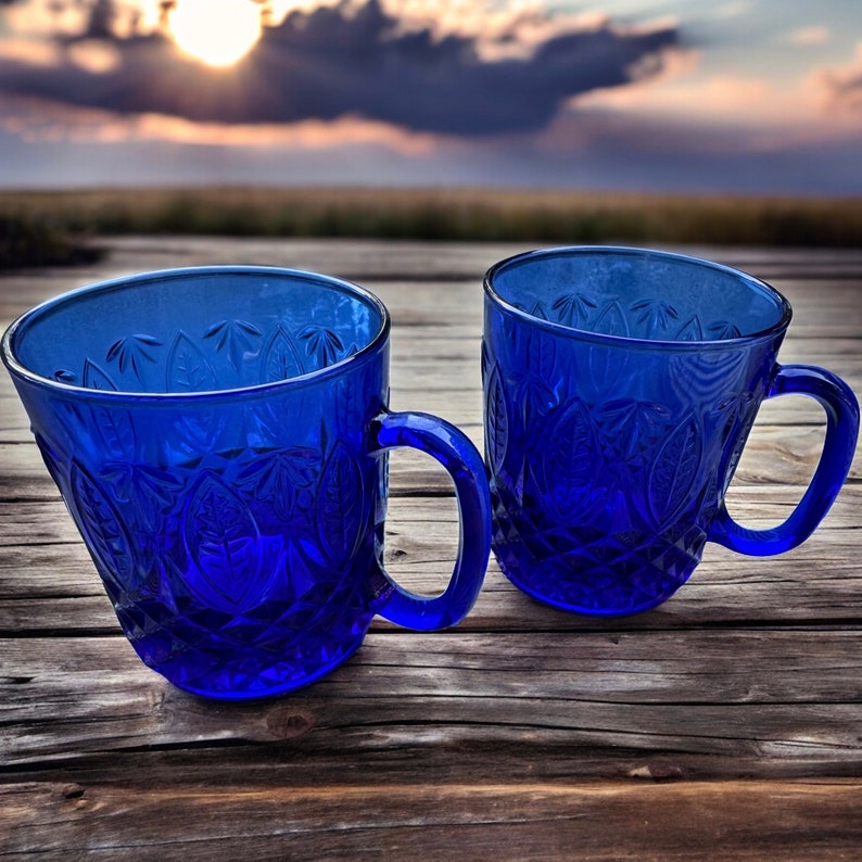May include: Two cobalt blue glass mugs with a textured pattern. The mugs have a handle on the right side and are sitting on a wooden surface.