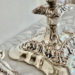 May include: A silver-toned metal candlestick base featuring an ornate, raised leaf-like relief pattern around the square pedestal. The surface has a reflective, polished finish with visible signs of aging and tarnishing along the decorative edges.