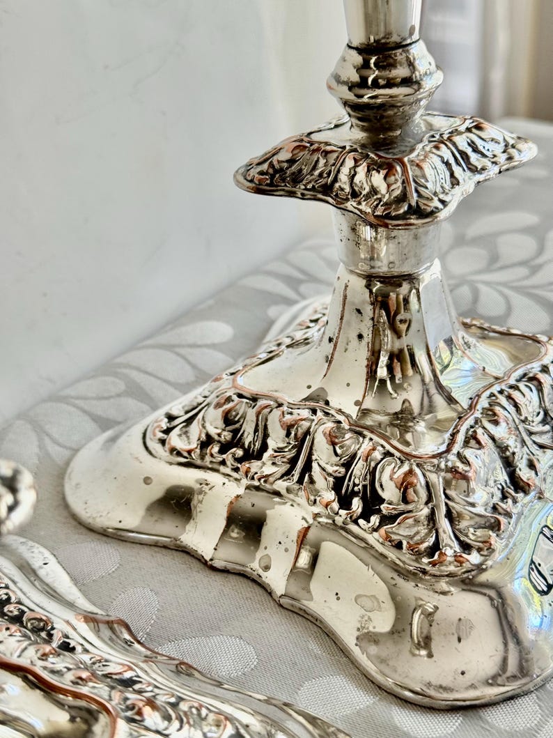 May include: A silver-toned metal candlestick base featuring an ornate, raised leaf-like relief pattern around the square pedestal. The surface has a reflective, polished finish with visible signs of aging and tarnishing along the decorative edges.