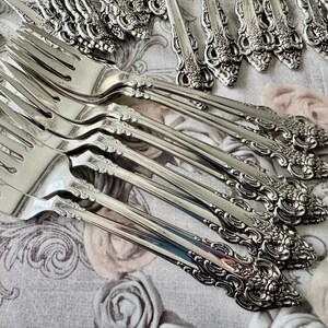 May include: Close-up of several ornate silver forks with detailed floral and scrollwork designs. The forks are arranged on a floral patterned fabric, showcasing their polished surfaces and intricate craftsmanship. A collection of silverware.