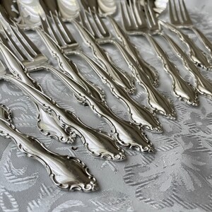 May include: A collection of ornate silver forks arranged on a patterned white tablecloth. The forks have a detailed, floral-like design along the handles and tines. The silverware is reflective, suggesting a polished finish.
