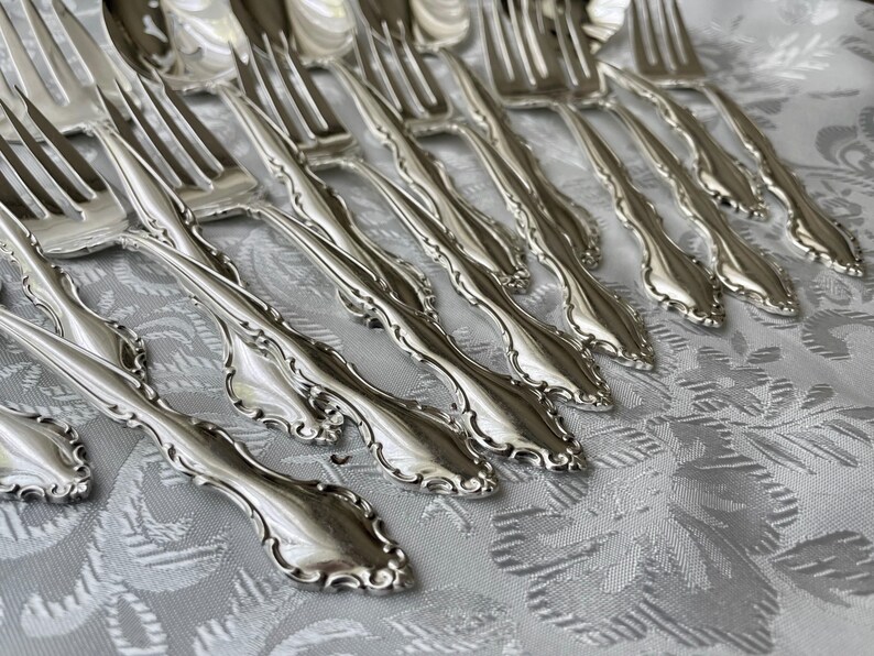 May include: A collection of ornate silver forks arranged on a patterned white tablecloth. The forks have a detailed, floral-like design along the handles and tines. The silverware is reflective, suggesting a polished finish.