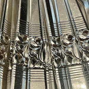 May include: A collection of silver-toned metal flatware handles arranged side-by-side. Each handle features a polished finish with a series of horizontal lines near the base and a raised, ornate, swirling floral pattern in the center.