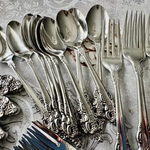 May include: An assortment of vintage silverware, including spoons, forks, and serving spoons. The silverware has ornate floral detailing on the handles and a shiny, reflective surface. The arrangement suggests a formal dining setting.