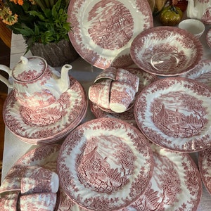 May include: A set of pink and white floral patterned china dinnerware, including plates, bowls, cups, and a teapot. The set features a cottage scene with trees and a fence.