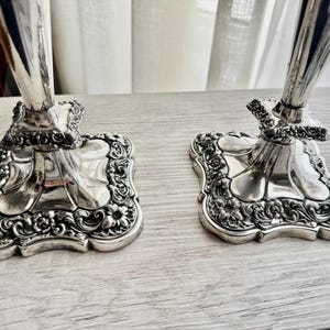 May include: A pair of silver-colored metal candlesticks featuring square bases with rounded corners. The base edges are decorated with embossed floral and scroll patterns, while the stems rise from a central pedestal with a smooth finish.
