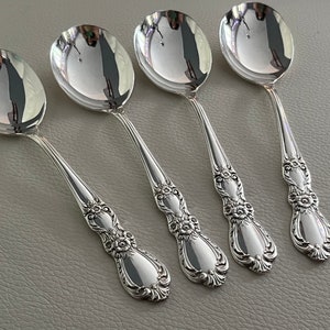 May include: A set of four polished silver-toned spoons featuring ornate floral and scrollwork detailing on the handles. Each spoon has a smooth, reflective oval bowl and a decorative, textured handle design that tapers toward the end.