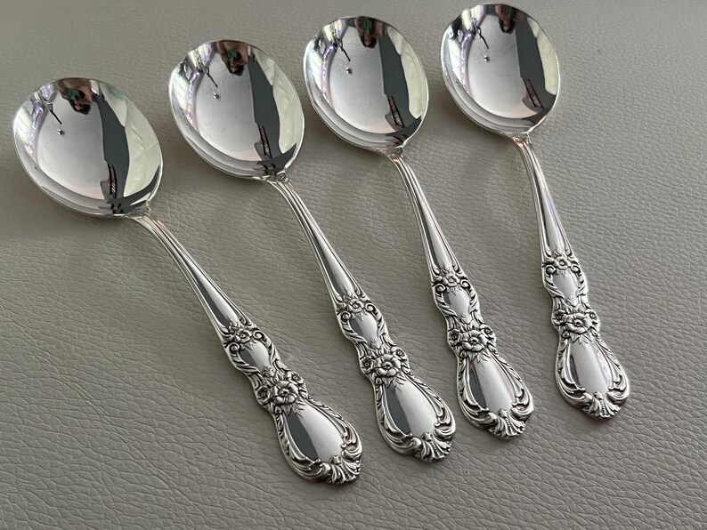 May include: A set of four polished silver-toned spoons featuring ornate floral and scrollwork detailing on the handles. Each spoon has a smooth, reflective oval bowl and a decorative, textured handle design that tapers toward the end.