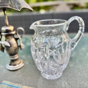 Puede incluir: Una jarra de crema de vidrio tallado transparente con asa y boquilla, que muestra patrones intrincados. Junto a ella, una pieza decorativa plateada con una pantalla de malla. Un mantel individual de bamb&uacute; con detalles azules en primer plano.