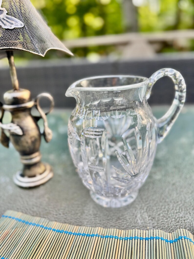Puede incluir: Una jarra de crema de vidrio tallado transparente con asa y boquilla, que muestra patrones intrincados. Junto a ella, una pieza decorativa plateada con una pantalla de malla. Un mantel individual de bamb&uacute; con detalles azules en primer plano.
