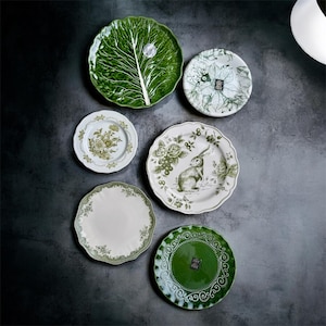 May include: A selection of decorative plates with diverse designs. One plate is shaped like a green cabbage leaf. Other plates feature floral and rabbit motifs in green and white. The plates are arranged on a dark grey surface.