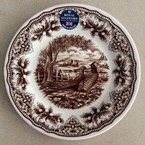 Op de afbeelding: Een witte keramische bord met een bruine transferprint van twee fazanten in een landschap met bomen en een hek. De rand is versierd met een herhalend patroon van eikenbladeren en eikels. Een blauwe Royal Stafford sticker bevindt zich aan de bovenkant.
