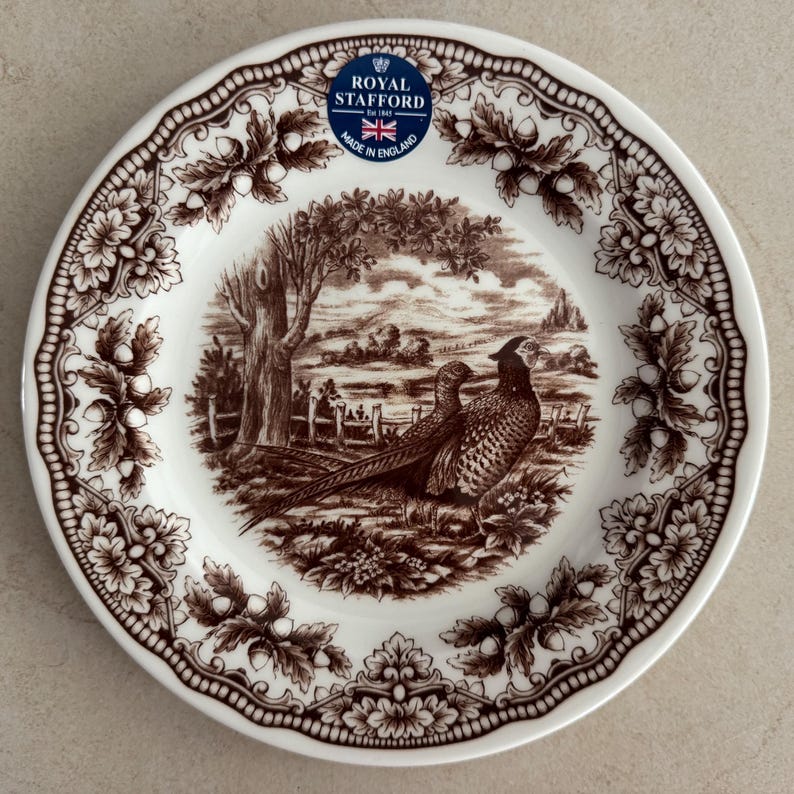 Op de afbeelding: Een witte keramische bord met een bruine transferprint van twee fazanten in een landschap met bomen en een hek. De rand is versierd met een herhalend patroon van eikenbladeren en eikels. Een blauwe Royal Stafford sticker bevindt zich aan de bovenkant.