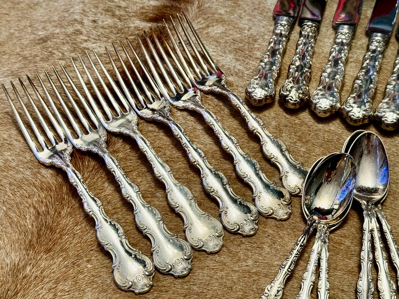 May include: A set of silver flatware, including forks and spoons, with ornate handles. The forks have a decorative design with a scalloped edge. The spoons have a similar design with a rounded bowl.