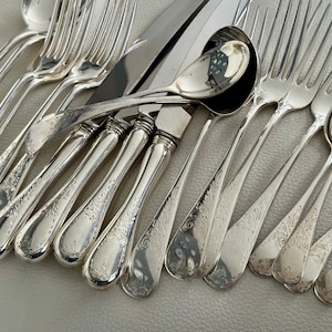 May include: An assortment of antique silverware, including forks, knives, and spoons, arranged on a light-colored surface. The silverware is made of a shiny, reflective metal, with ornate floral patterns etched into the handles. The collection includes various sizes of spoons and forks.