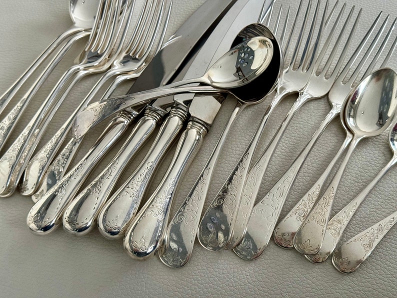 May include: An assortment of antique silverware, including forks, knives, and spoons, arranged on a light-colored surface. The silverware is made of a shiny, reflective metal, with ornate floral patterns etched into the handles. The collection includes various sizes of spoons and forks.