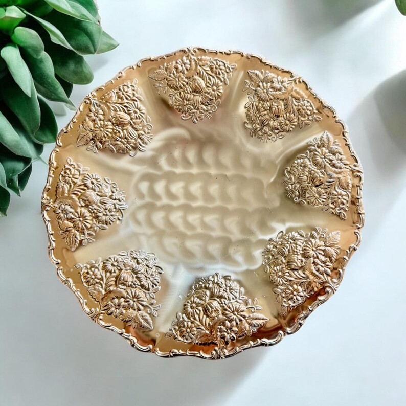 May include: A gold-toned, decorative serving plate with a scalloped edge. The plate features embossed floral designs around the rim and in the center. The center has a textured, ridged pattern.
