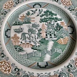 May include: A white ceramic plate with a detailed green and brown Asian-inspired landscape design. The plate features pagodas, trees, a boat, and a bridge, surrounded by a decorative border. The plate's design is intricate and detailed.