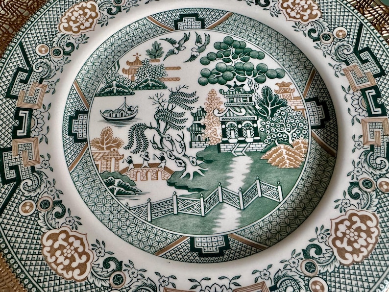 May include: A white ceramic plate with a detailed green and brown Asian-inspired landscape design. The plate features pagodas, trees, a boat, and a bridge, surrounded by a decorative border. The plate's design is intricate and detailed.